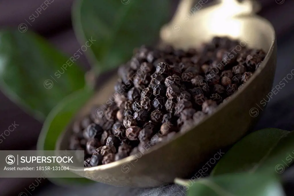 Brass spoon with black peppercorns and leaves on textile, close up