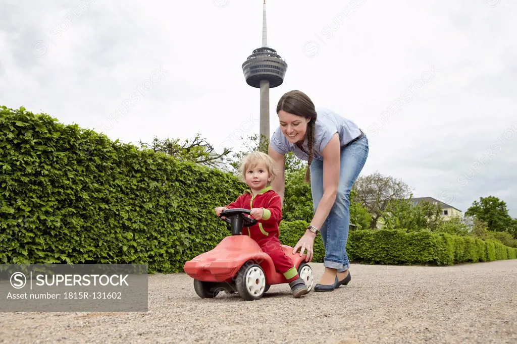 Germany, Cologne, Mother carrying daughter in toy car, smiling