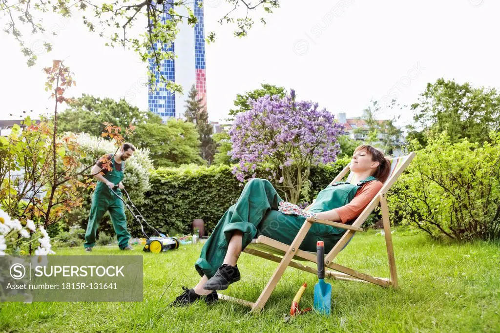 Germany, Cologne, Young woman relaxing on deck chair while man mowing lawn