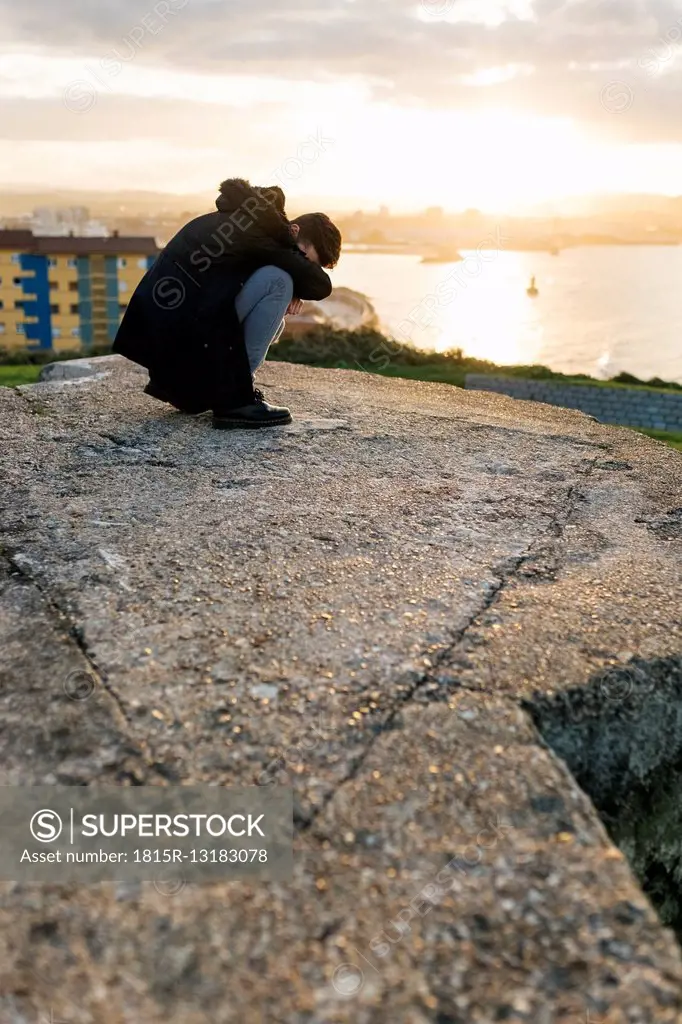 Spain, Gijon, man crouching on a wall at evening twilight covering face with his arm