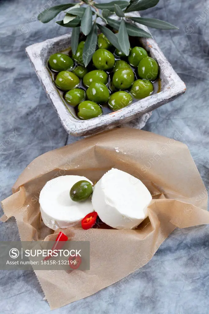 Goat cheese with green olives in olive oil and chili on brown paper, close up