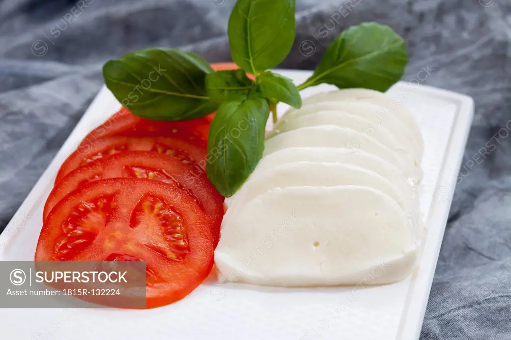 Slices of mozzarella cheese, tomatoes and basil herb on chopping board, close up