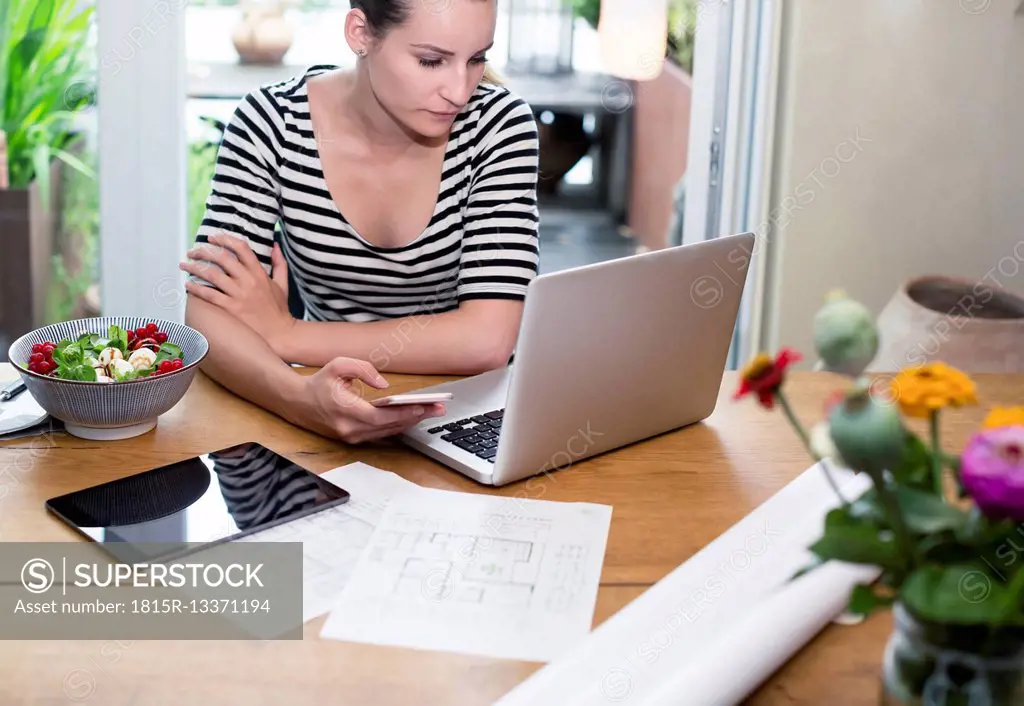 Woman at desk using laptop and cell phone next to construction plan and salad