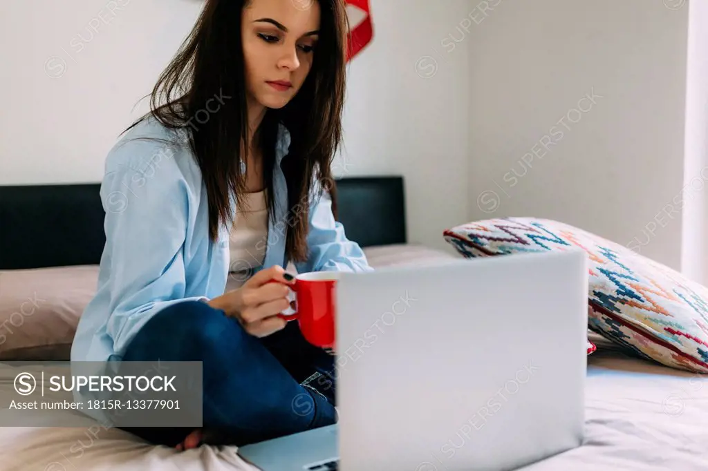 Young woman sitting on bed, coffee cup, laptop and smartphone