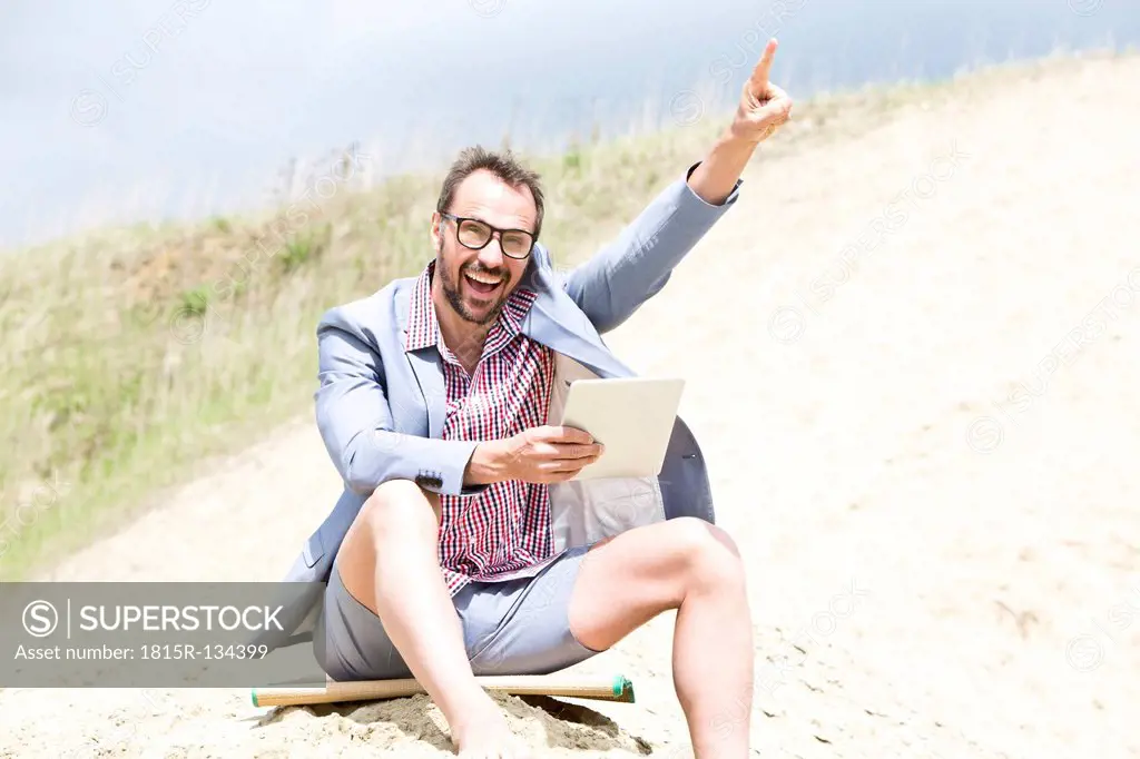 Germany, Bavaria, Portrait of businessman sitting on sand with digital tablet, smiling