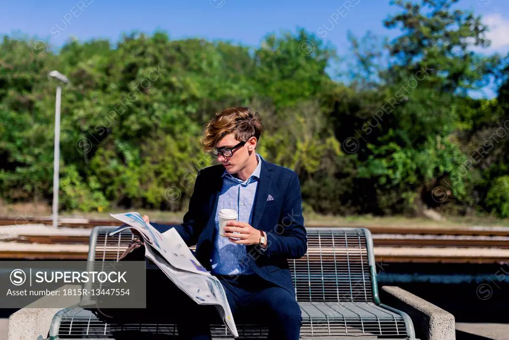 Young businessman with coffee to go sitting on bench at platform reading newspaper