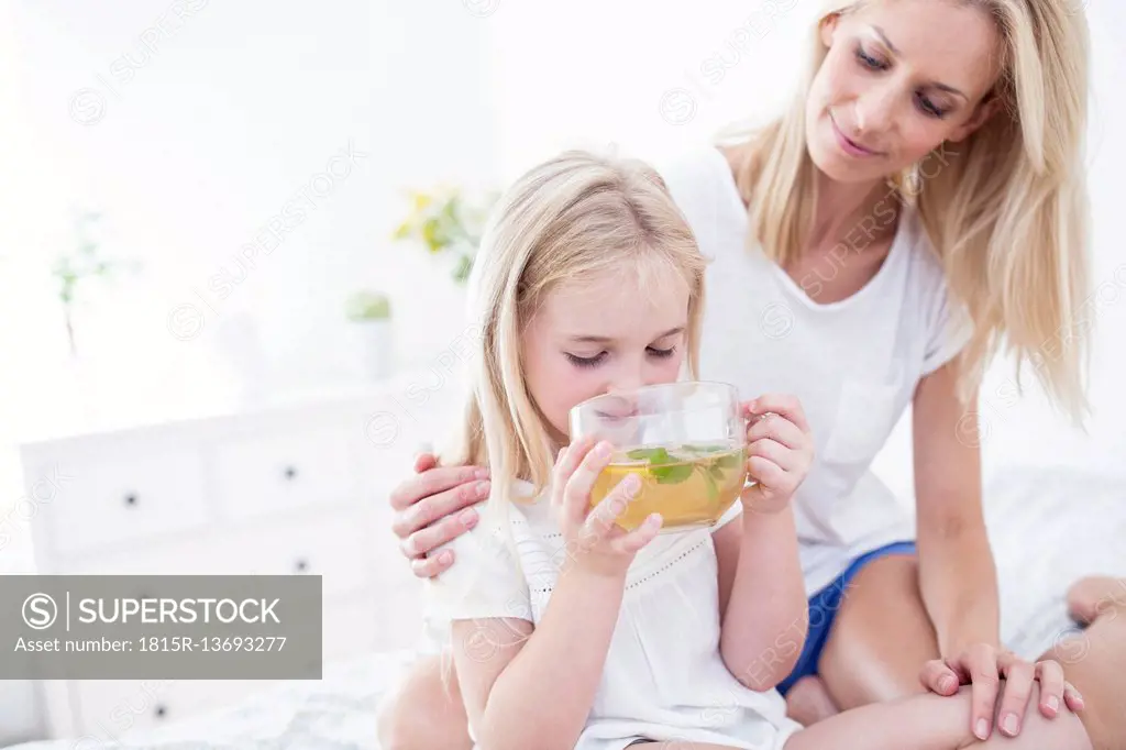 Mother watching daughter drinking cup of tea on bed