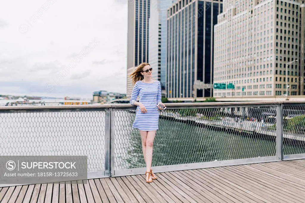 USA, New York City, Young woman standing in Manhattan at railing, holding smart phone