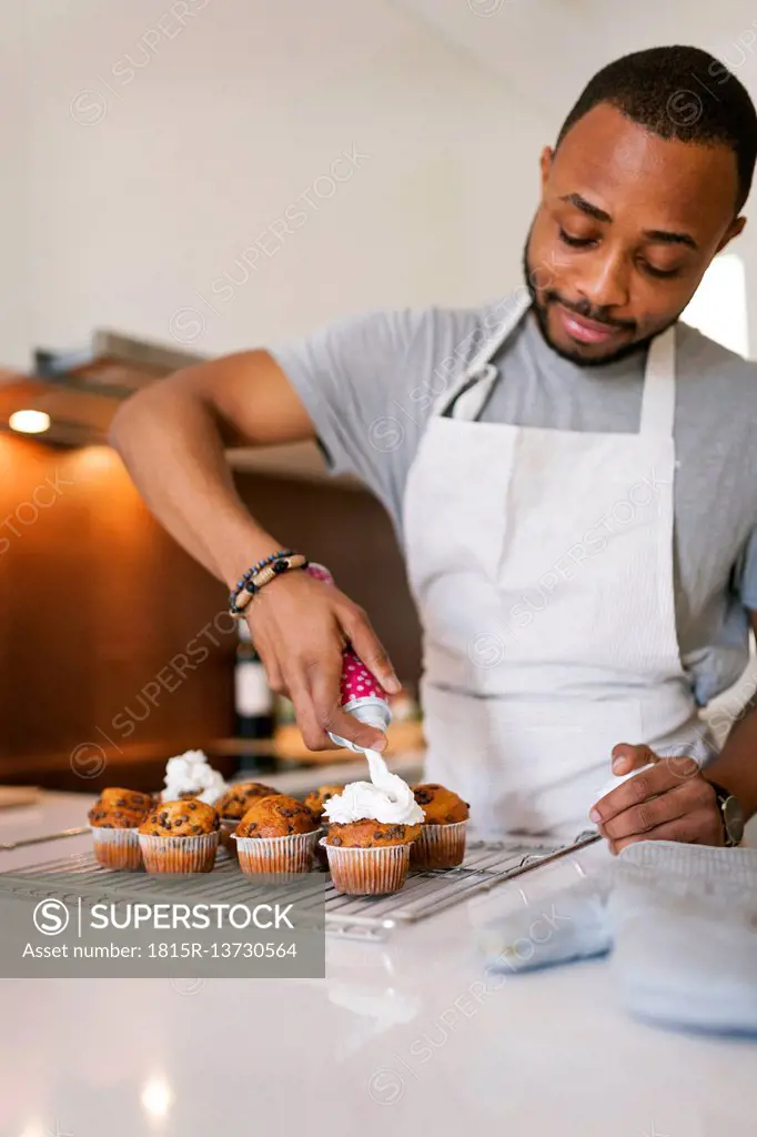 Young man baking cup cakes at home, topping them with cream