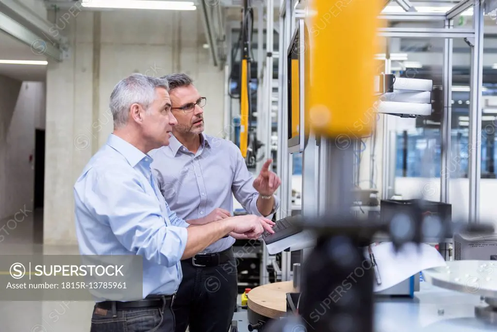 Two businessmen in modern factory hall talking at computer monitor