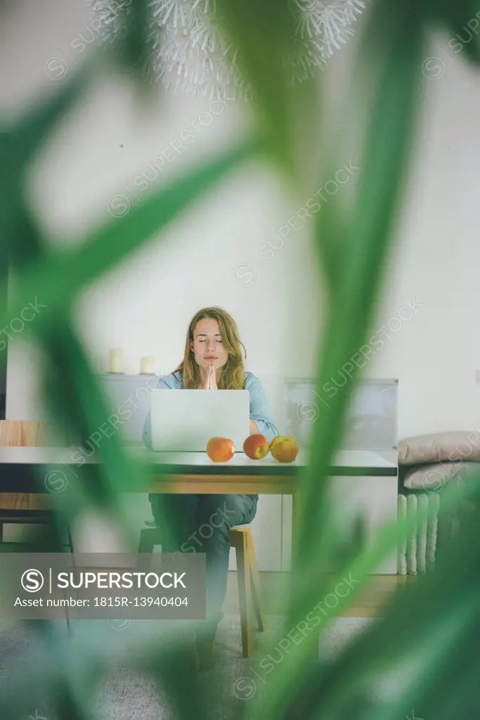 Young woman with closed eyes sitting at table with apples and laptop