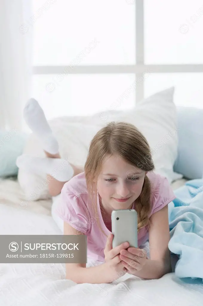 Germany, Bavaria, Girl using smart phone on bed, smiling