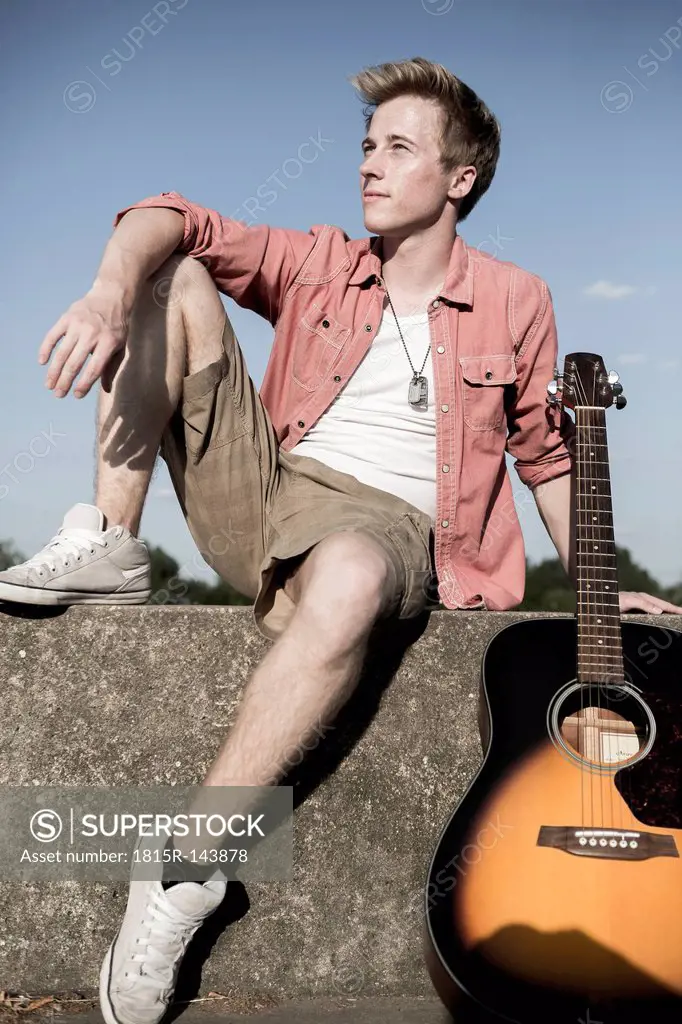 Germany, Young man sitting on wall with guitar