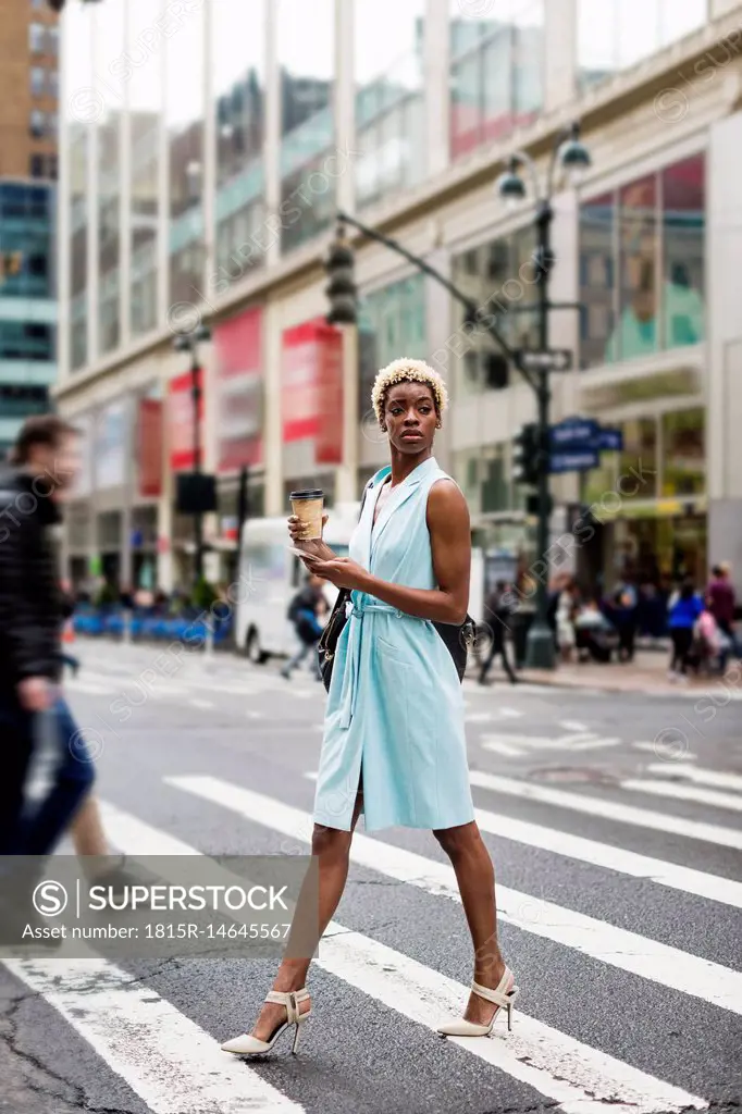 USA, New York, young blonde african-american woman with cup of coffee and smart phone crossing street