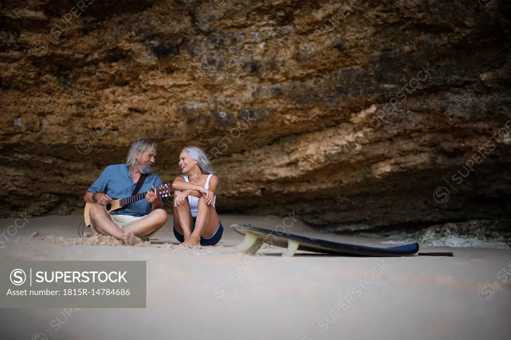 Handsome senior couple sitting on the beach, man playing guitar