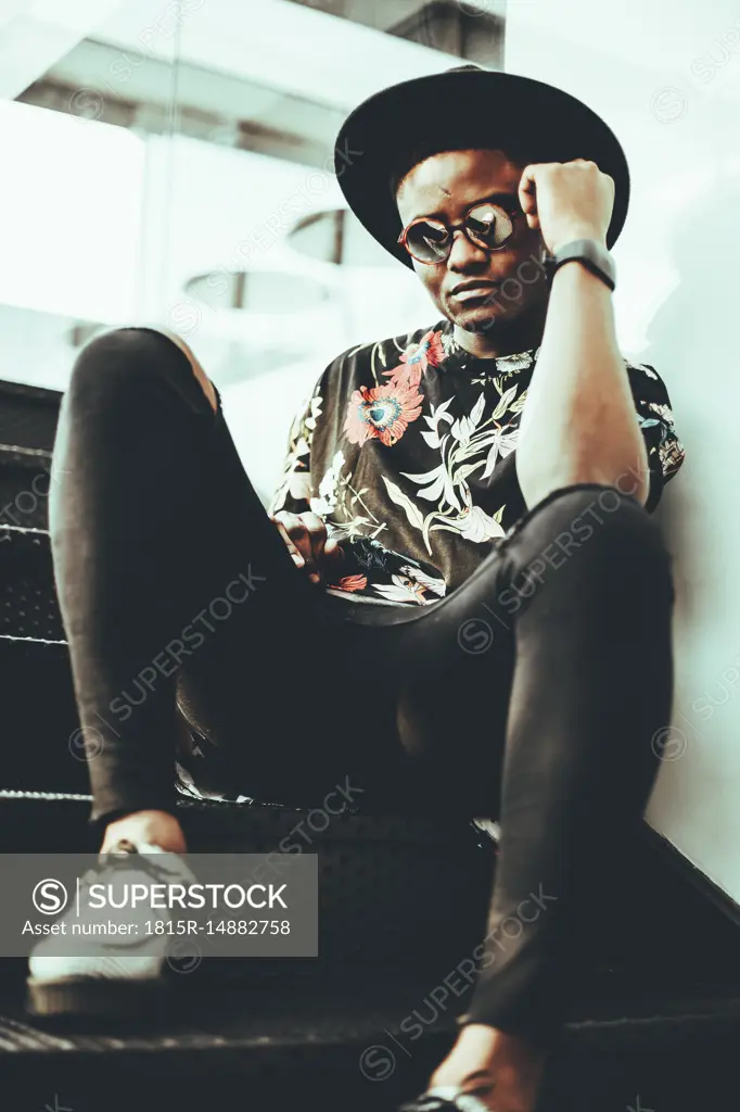 Portrait of fashionable man wearing hat, sunglasses and black t-shirt with floral design siitng on stairs
