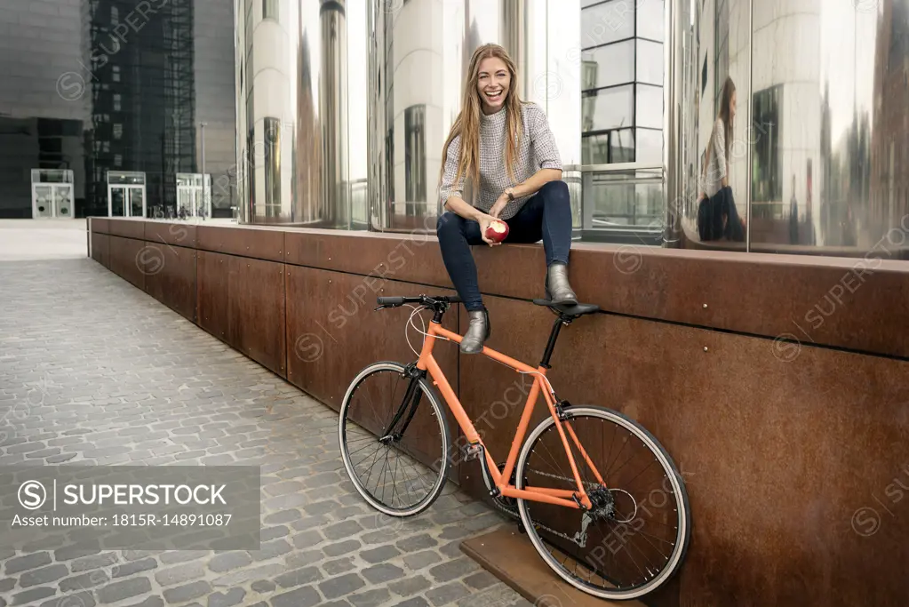 Happy young woman with bicycle having a break in the city eating an apple