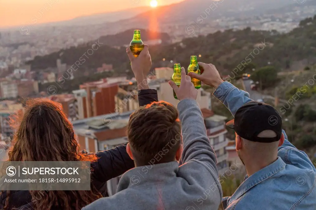Spain, Barcelona, three friends with beer bottles on a hill overlooking the city at sunset