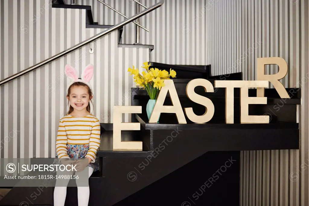 Portrait of smiling girl with bunny ears sitting on stairs next to word 'Easter'