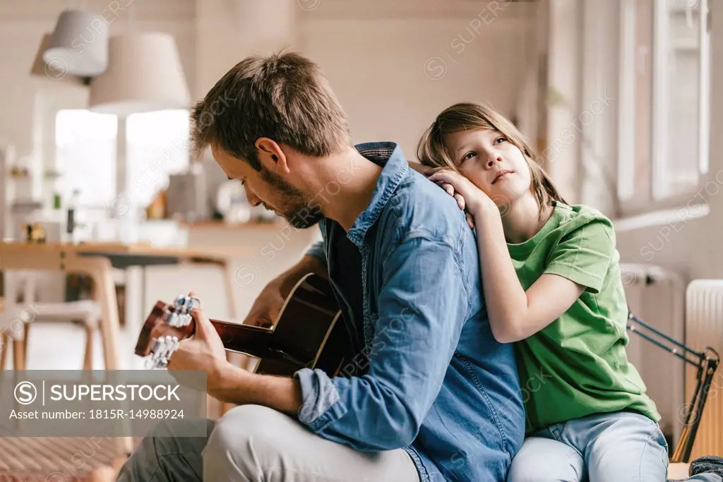 Son leaning against father playing guitar at home