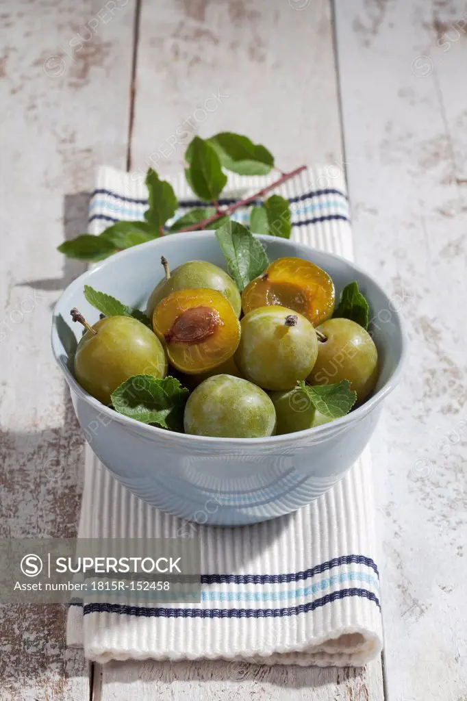 Sliced and whole greengages (Prunus domestica subsp. italica var. claudiana) in a bowl on white wooden table, studio shot