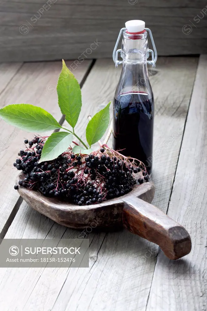Elderberries (Sambucus), leaves on a wooden shovel and a bottle of elderberry juice on white wooden table, studio shot