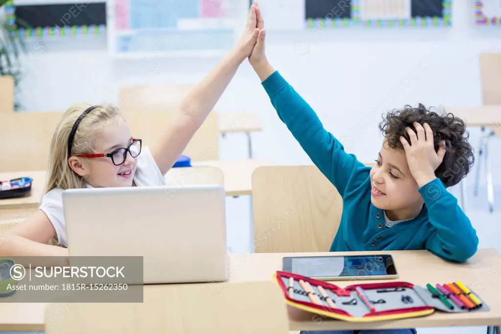 Schoolboy and schoolgirl with laptop high fiving in class