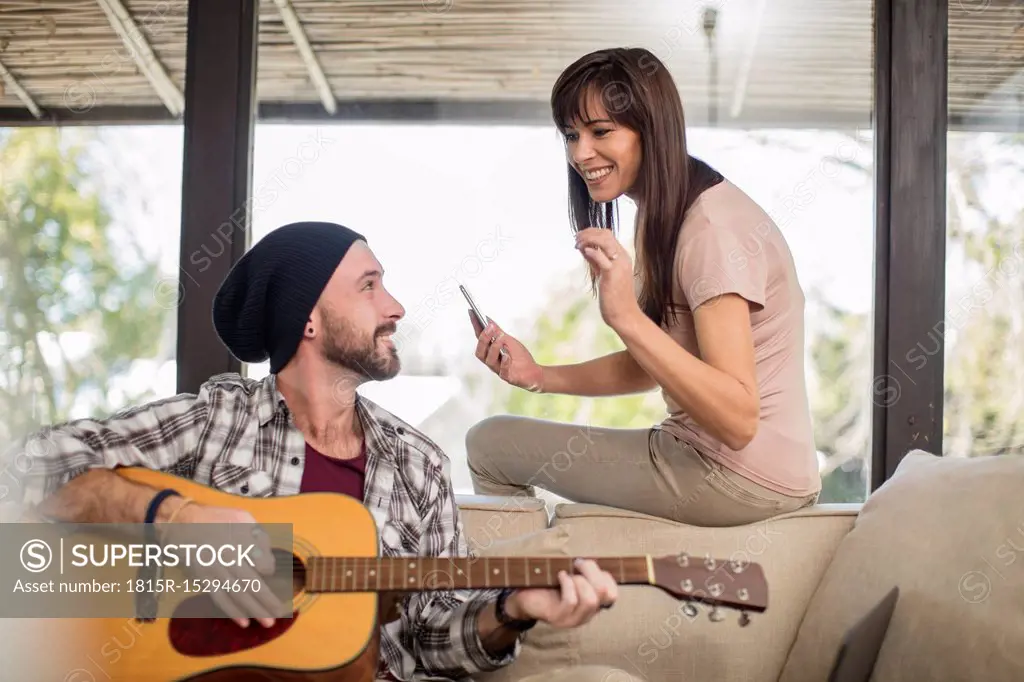 Young man at home sitting on couch playing guitar for woman