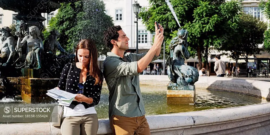 Portugal, Lisboa, Baixa, Rossio, Praca Dom Pedro IV, young couple with city map and smart phone in front of a fountain
