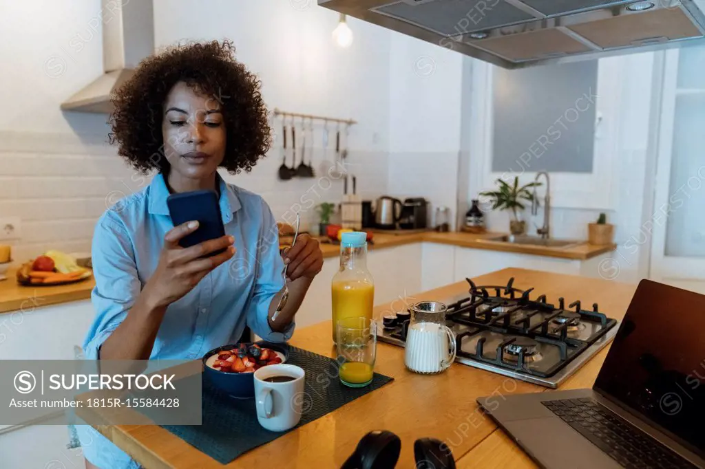 Woman having breakfast in her kitchen, using smartphone