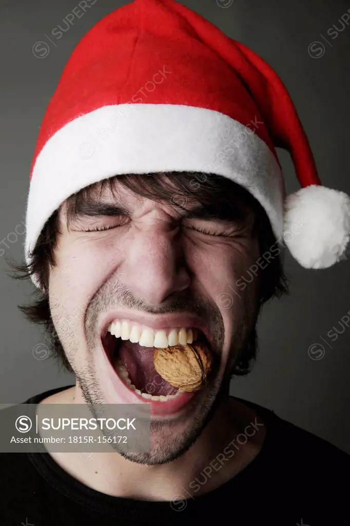 Young man with Santa hat trying to crack a walnut with his teeth, studio shot
