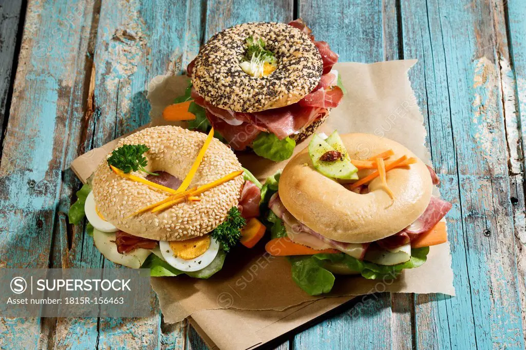 Three different bagels garnished with salami, sausage, slices of bacon, rocket salad, lettuce, cucumber carrot, egg, cream cheese and cress and parsle...