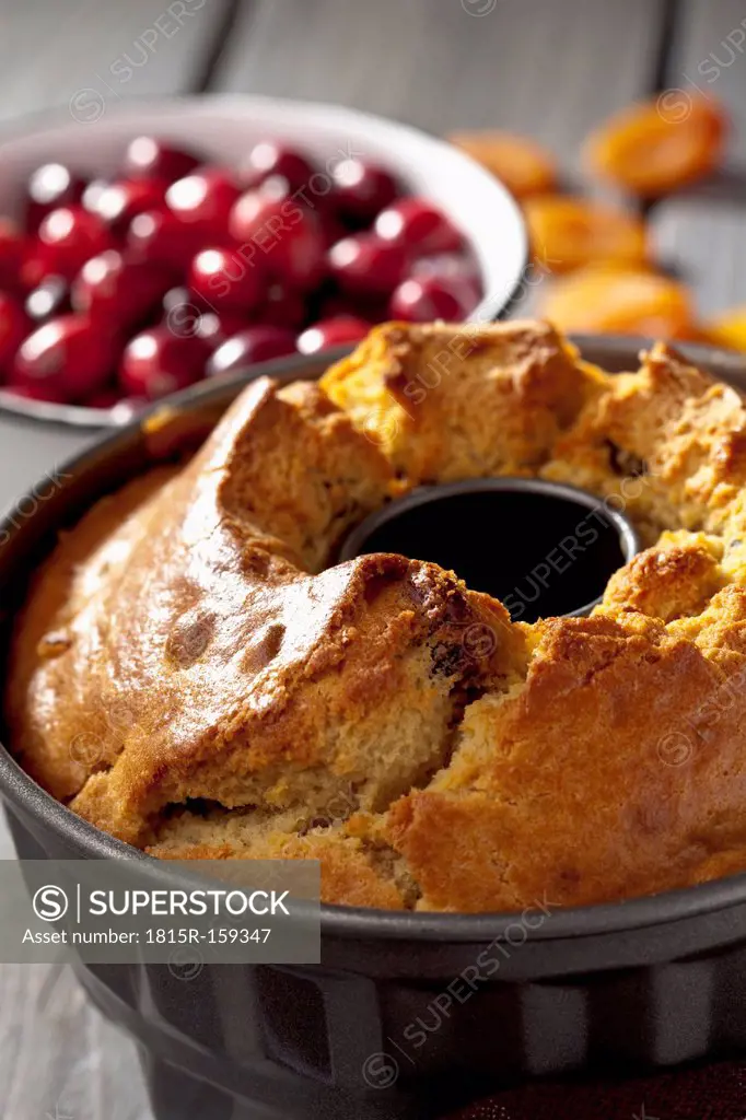 Cake pan with baked ring cake and a bowl of cranberries on grey wooden table