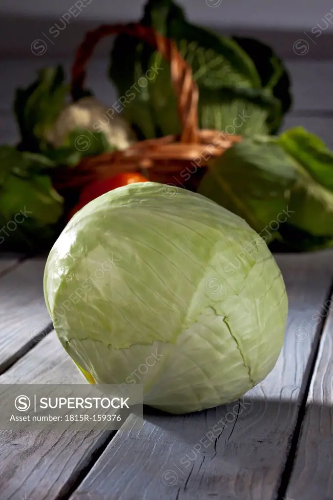 White cabbage (Brassica oleracea convar. capitata var. alba) and basket with other vegetables at background on grey wooden table