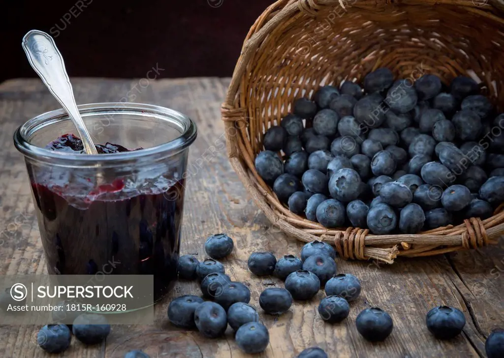 Wickerbasket with blueberries (Vaccinium myrtillus) and glass of blueberry jam on wooden table