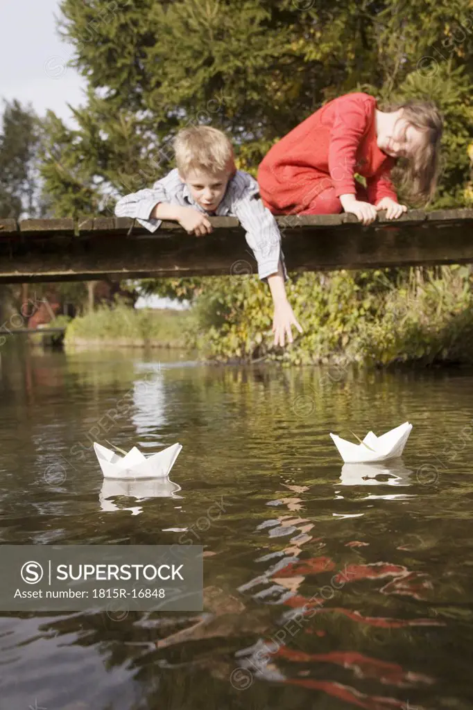 Boy (10-12) and girl (7-9) on bridge, watching paper boats in water