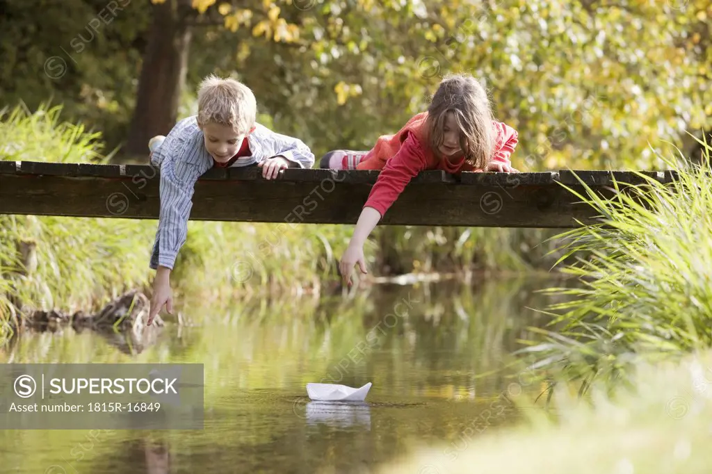 Boy (10-13) and girl (7-9) lying on bridge, watching paper boats in water