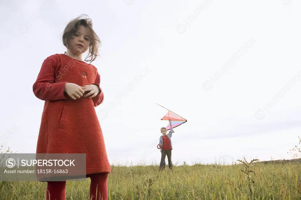 Girl (7-9) standing in meadow, boy (10-12) holding kite in background