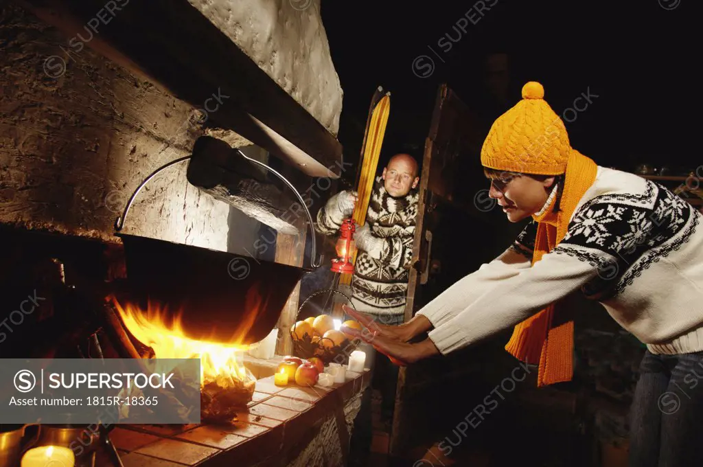 Mid adult couple in alpine hut, woman warming hands at fireplace