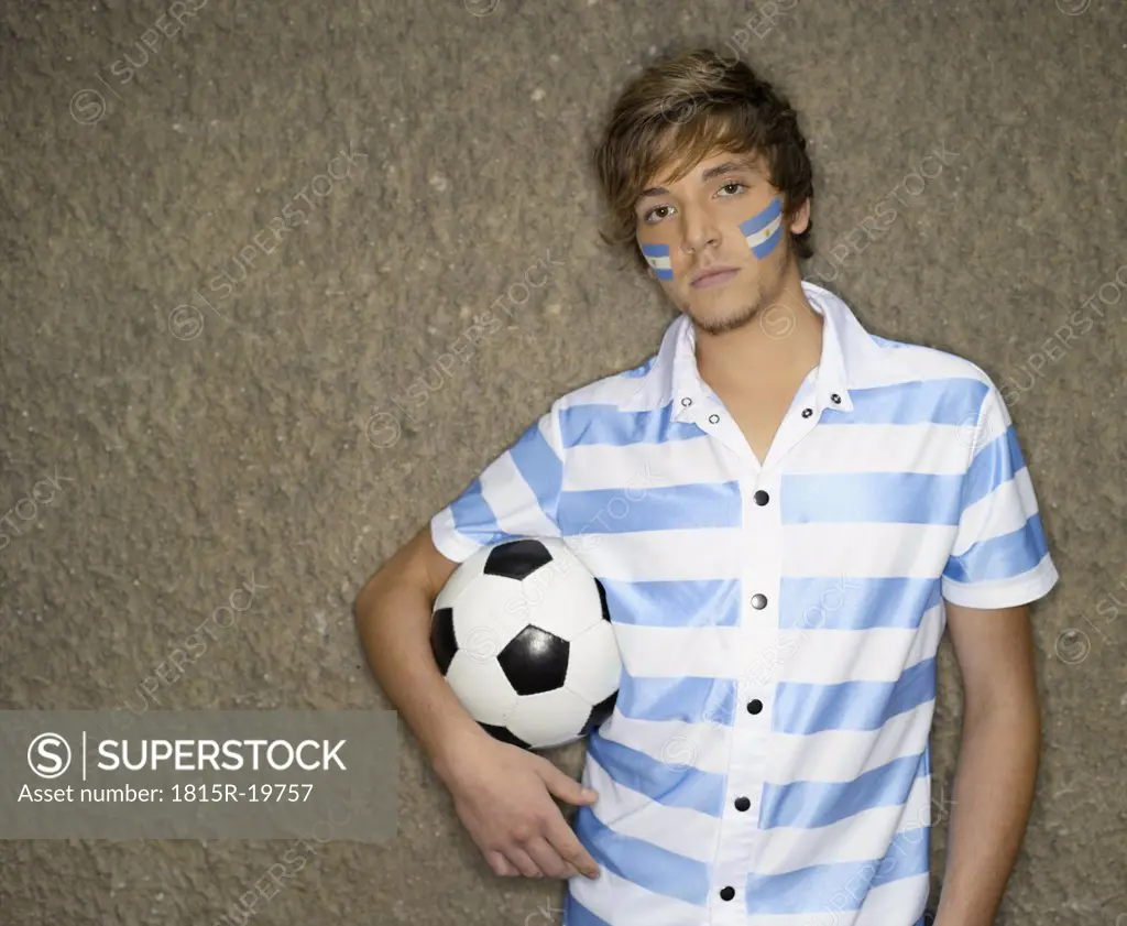 Young man with Argentinian flag painted in his face, and with soccer ball leaning against wall, portrait