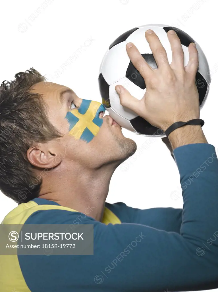 Young man with Sweden flag painted on face, kissing soccer ball, side view