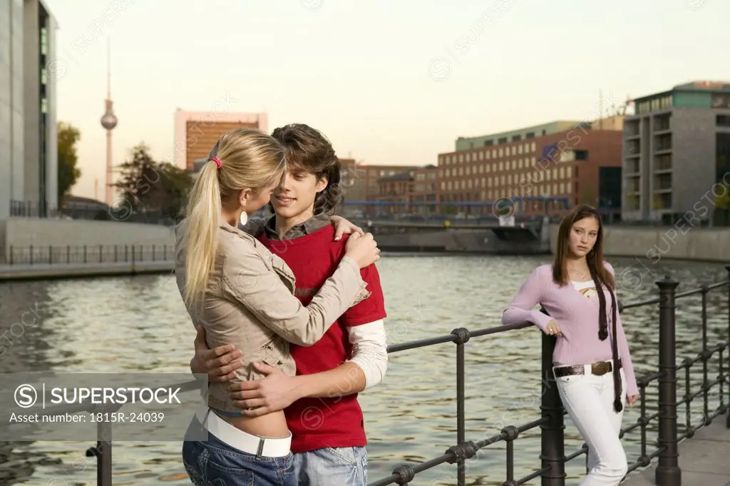 Couple embracing at river, young woman standing in background