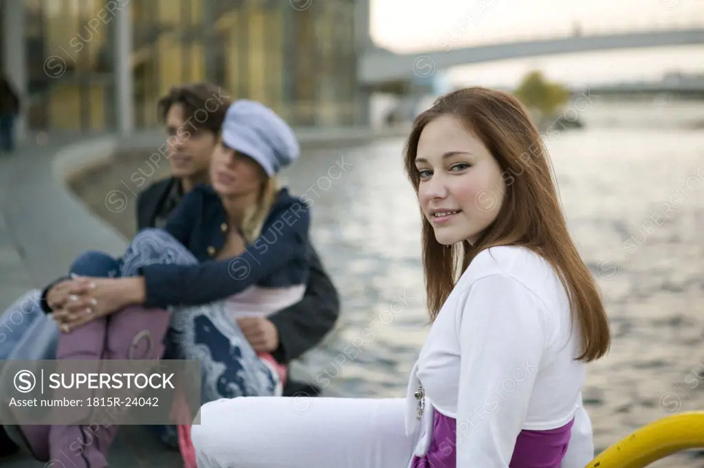 Young woman sitting at river, couple in background