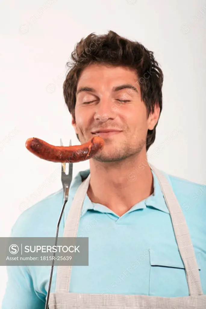 Young man smelling grilled sausage on fork, eyes closed, close-up