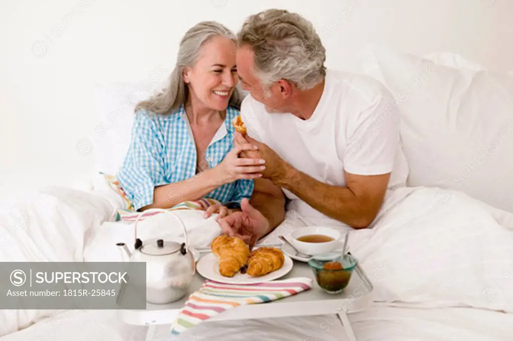 Mature couple sitting on bed with breakfast, smiling