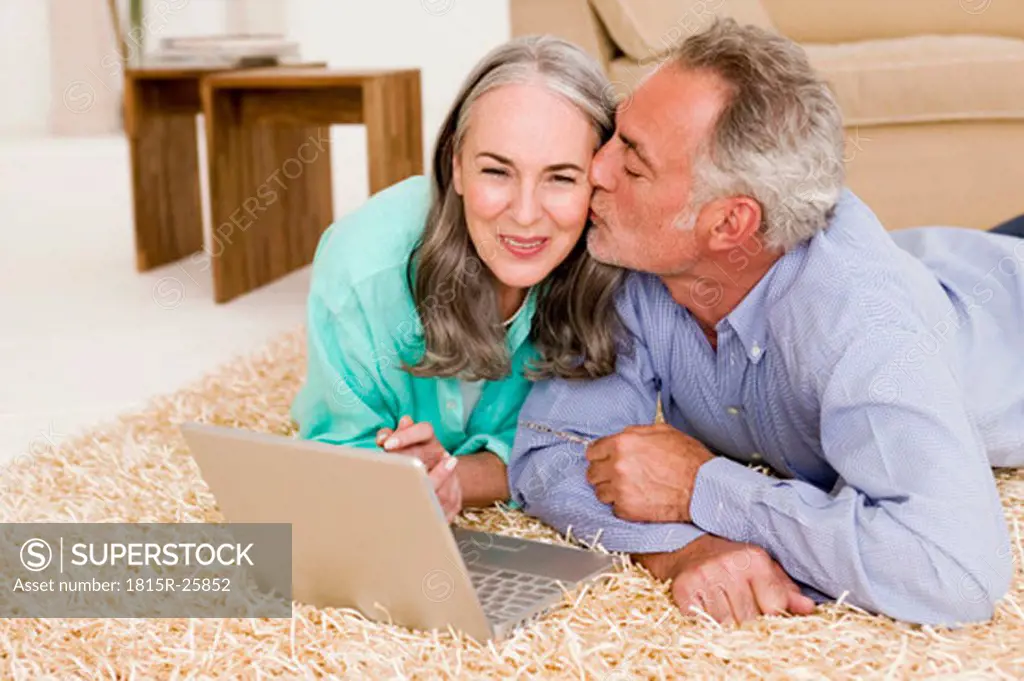 Mature couple lying on carpet with laptop, man kissing woman