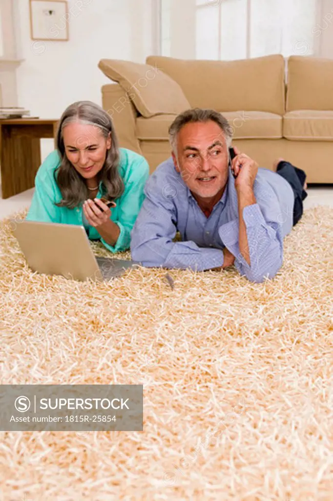 Mature couple lying on carpet with laptop, man using mobile phone