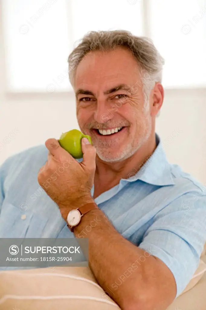 Mature man sitting on sofa holding apple, smiling, portrait