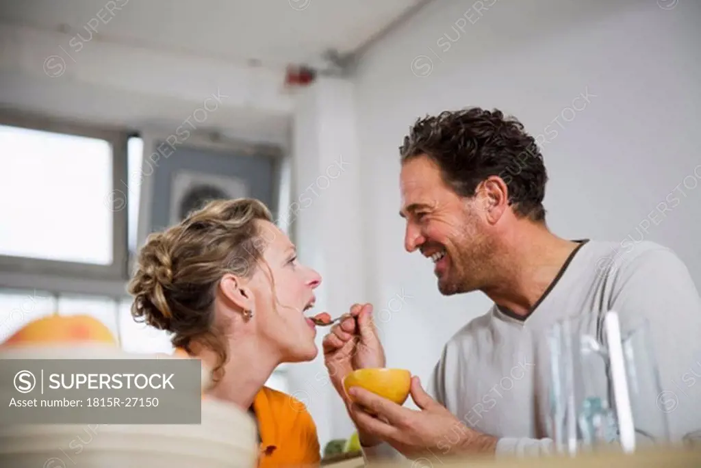 Mature man feeding grapefruit to woman in kitchen, smiling, low angle view