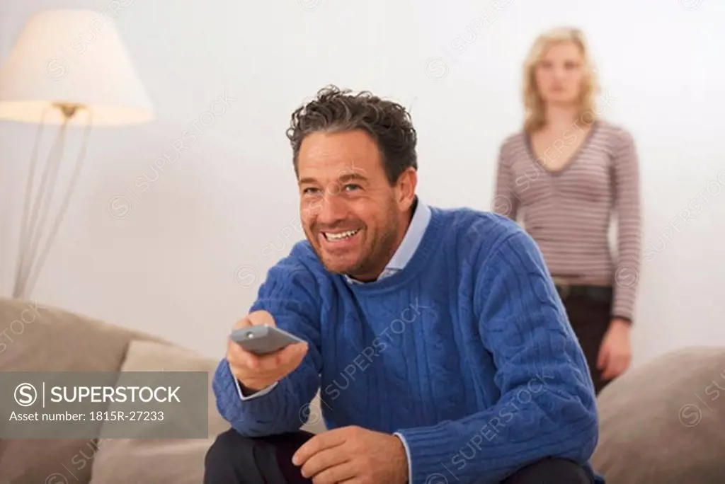 Man sitting on sofa, holding remote control, daughter in background